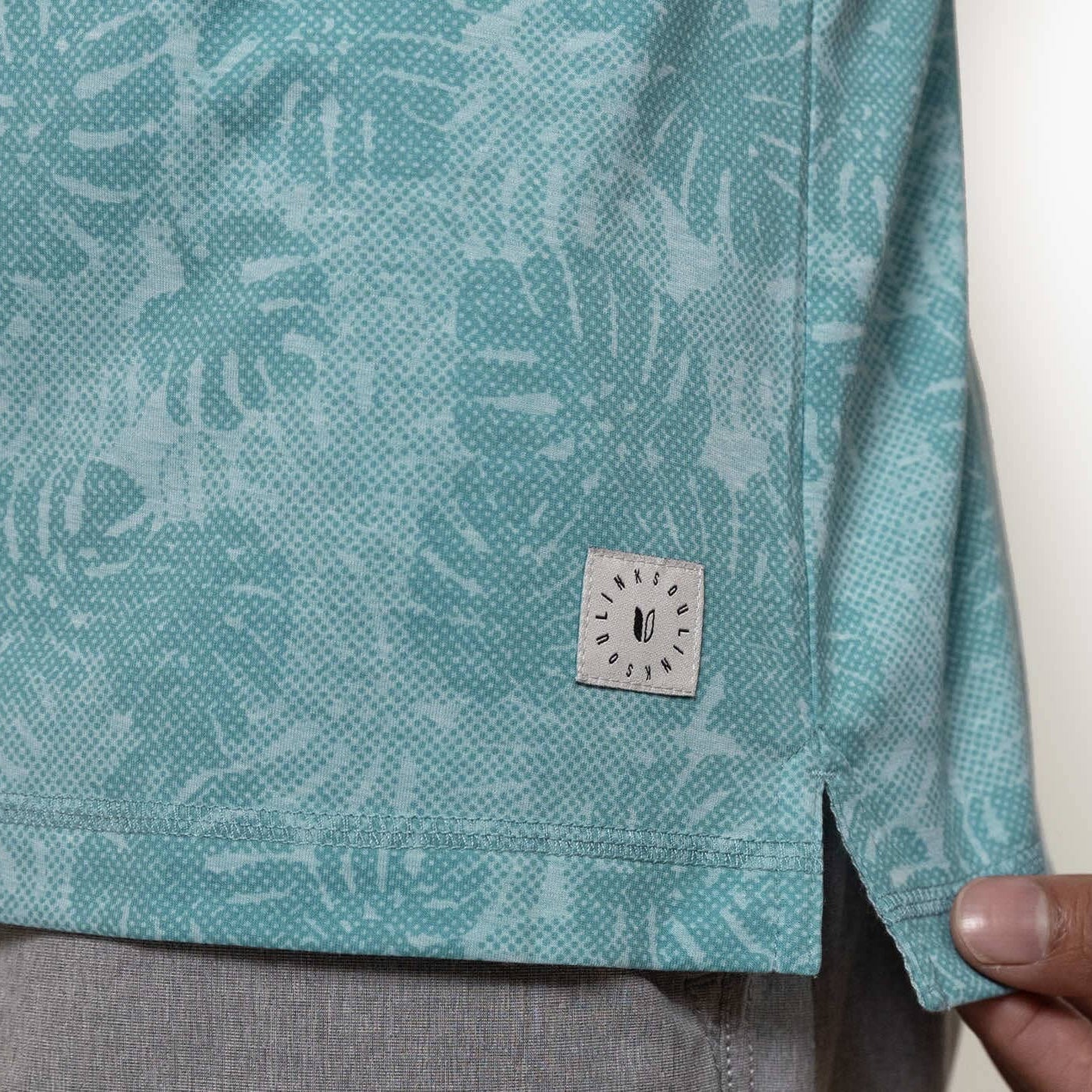 Teal patterned shirt with a brand label held by a hand against a neutral background