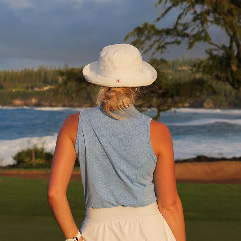 Person wearing a white hat and blue sleeveless top standing by a scenic ocean view.